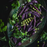 Purple pea pods and green pea shoots arranged densely on a dark ceramic plate